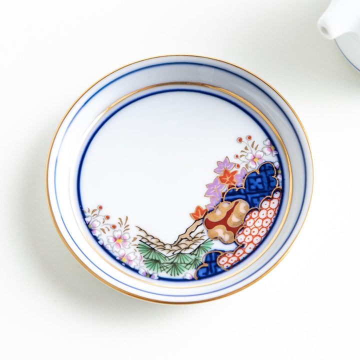 A small white sauce dispenser with intricate traditional Japanese patterns in blue, red, and gold, accompanied by a matching saucer.