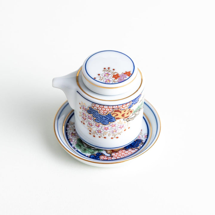 A small white sauce dispenser with intricate traditional Japanese patterns in blue, red, and gold, accompanied by a matching saucer.