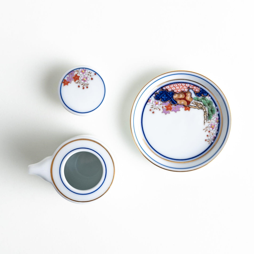 A small white sauce dispenser with intricate traditional Japanese patterns in blue, red, and gold, accompanied by a matching saucer.