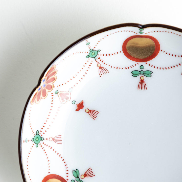 A round white sauce dish with delicate floral and ribbon patterns in gold, green, and red, accented by small scalloped edges.