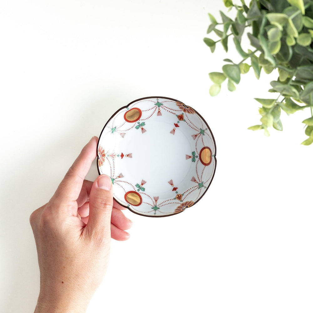 A round white sauce dish with delicate floral and ribbon patterns in gold, green, and red, accented by small scalloped edges.