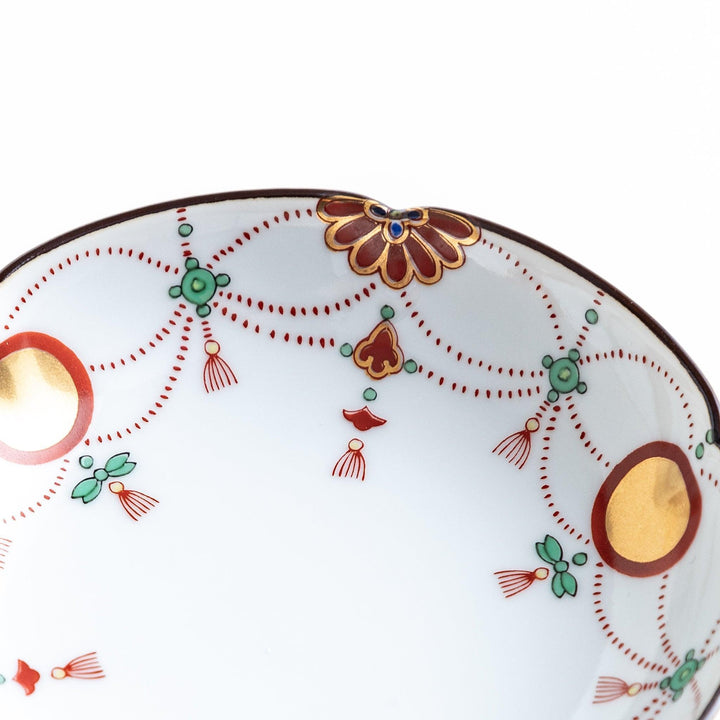 A round white sauce dish with delicate floral and ribbon patterns in gold, green, and red, accented by small scalloped edges.