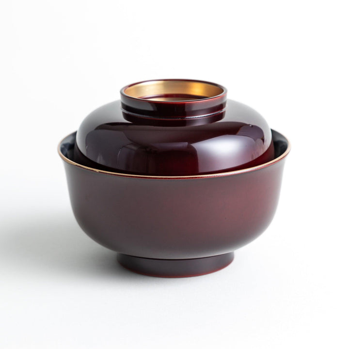 A glossy lacquer bowl with a matching lid, featuring a gold rim around the edge, ideal for serving soups or stews. Available in black or red.