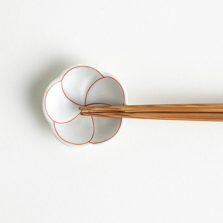 A small, white flower-shaped chopstick rest with a minimalist red swirl pattern.