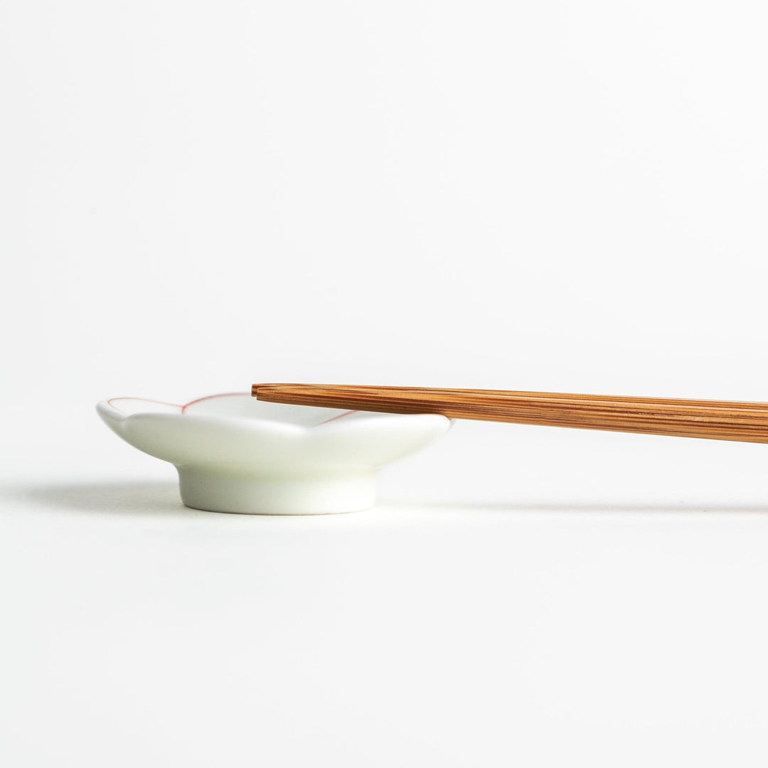 A small, white flower-shaped chopstick rest with a minimalist red swirl pattern.