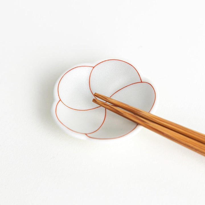 A small, white flower-shaped chopstick rest with a minimalist red swirl pattern.